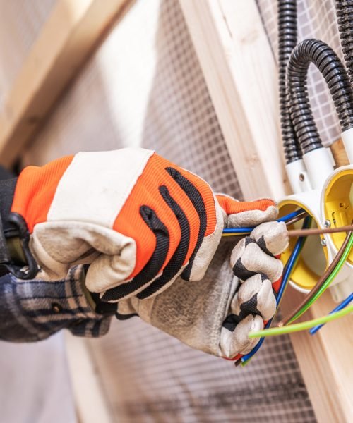 An electrician connects colored wires to a junction box while wearing gloves and working diligently. The setting shows a construction site with wooden frames.