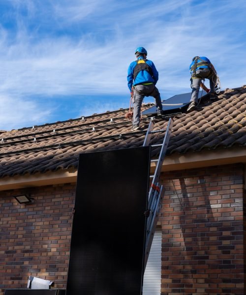 Two technicians installing photovoltaic panels on a roof, using a ladder to carry the panels up